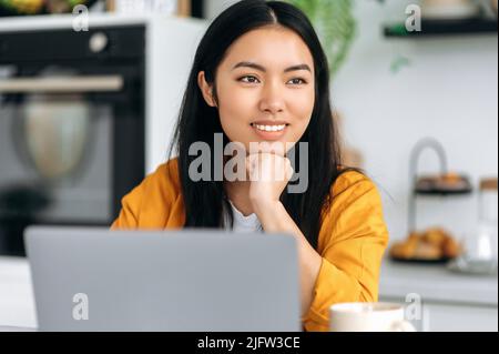 Ritratto di una bella donna bruna asiatica, giovane e affascinante, freelance, designer, seduto sul posto di lavoro con un laptop, guardando via, pensando, sognando, progettando il futuro, sorridendo felice Foto Stock