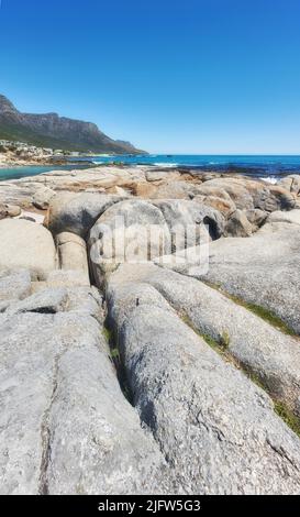 Paesaggio roccioso che conduce al mare sotto cielo blu nuvoloso copiare spazio a Camps Bay Beach, Città del Capo, Sudafrica. Massi sulla costa costiera con il Foto Stock