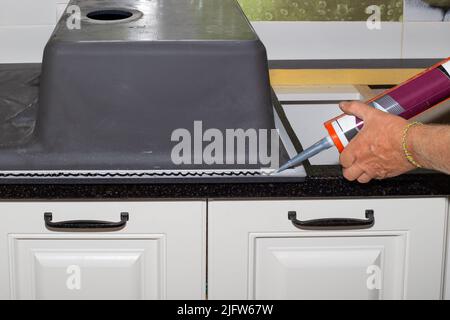 Applicazione di sigillante sanitario nero con una pistola per unghie al bordo del lavello della cucina. Installazione di un lavandino su un piano di lavoro della cucina. Foto Stock