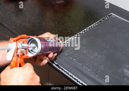 Applicazione di sigillante sanitario nero con una pistola per unghie al bordo del lavello della cucina. Installazione di un lavandino su un piano di lavoro della cucina. Foto Stock