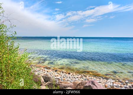 CopySpace al mare con sfondo cielo blu nuvoloso. Calme onde oceaniche che si lavano su pietre a una spiaggia vuota con barche a vela che navigano nel Foto Stock