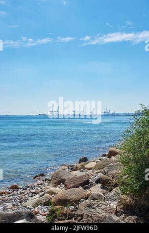 Copia lo spazio al mare con un cielo blu sfondo e gru porto in un porto all'orizzonte. Calme onde oceaniche che si lavano su una costa rocciosa. Panoramica Foto Stock