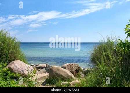 Una costa rocciosa poco profonda in una tranquilla giornata sulla spiaggia durante l'estate con erba che cresce sulla riva. Vista panoramica di un oceano blu cristallo con blu chiaro Foto Stock