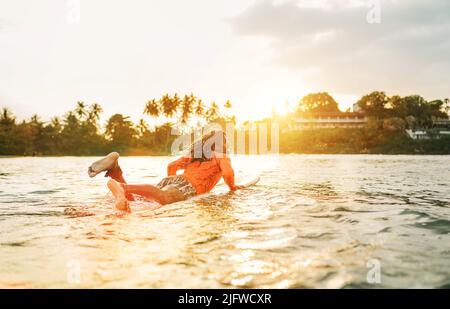 Uomo nero dai capelli lunghi che si paddling su una lunga tavola da surf al punto di surf nell'oceano Indiano. Palmeto ha littato raggi del tramonto sullo sfondo. Acqua estrema s Foto Stock