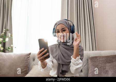 Donna musulmana seduta sul divano e videoconferenza chiamata sul cellulare. Videochiamata musulmana asiatica. Foto Stock
