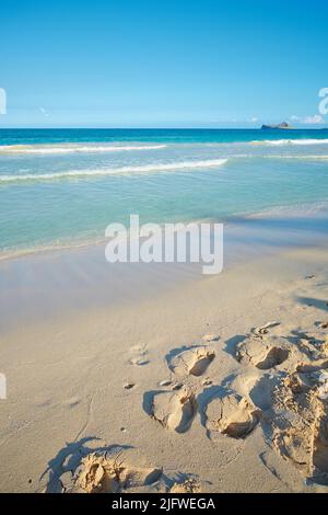 Le onde si lavano sulla spiaggia sabbiosa con le impronte di un resort tropicale ed esotico con il cielo blu chiaro sfondo e copyspace. Estate rilassante Foto Stock