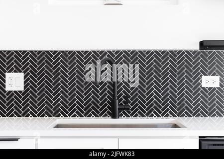 Un dettaglio lavandino della cucina girato con un rubinetto nero, marmo nero herringbone backsplash, e un piano di granito bianco. Foto Stock