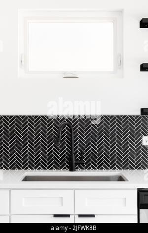 Un dettaglio lavandino della cucina girato con un rubinetto nero, marmo nero herringbone backsplash, e un piano di granito bianco. Foto Stock