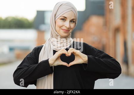 Bella giovane musulmana sorridente fiduciosa, entusiasta e allegra con le mani segno di amore, affetto, felice, sul petto che cammina sullo sfondo della città. Foto Stock