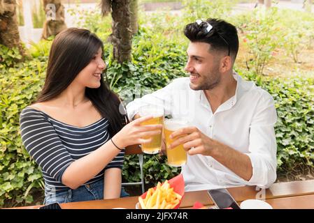 Romantica coppia giovane tostatura con birra fredda all'aperto in un parco o giardino sorridente e felice come celebrare il loro amore Foto Stock