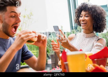 La giovane coppia si diverse a mangiare fast food al tavolo del ristorante ridendo mentre legge i media sul proprio cellulare Foto Stock
