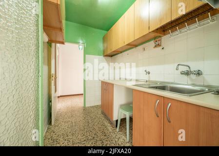 Angolo di una vecchia cucina con mobili in legno, piani bianchi, pavimenti in terrazzo e accesso ad una terrazza chiusa e soffitti dipinti di verde Foto Stock