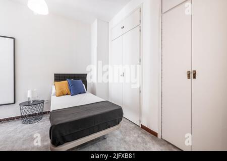 camera da letto con letto singolo, comodino in metallo e legno nero, specchio appeso alla parete e porte in legno bianco su diversi armadi a muro Foto Stock