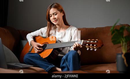 Attraente bella donna millenaria asiatica si rilassa seduto in salotto, suonando la sua chitarra acustica. Hobby o concetto di tempo libero Foto Stock