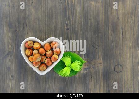 Nocciole fresche non sgusciate in un piatto a forma di cuore con foglie verdi in un piano in alto stendere la vita su un tavolo di legno con copyspace Foto Stock