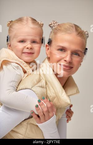 Tecnologia virtuale. Felice felice bella ragazza sorridente e guardando la macchina fotografica mentre la madre tiene in mano le sue braccia indossando occhiali digitali virtuali su biegio Foto Stock