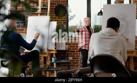 Insegnante senior che dà lezioni d'arte a diversi gruppi di studenti in un workshop scolastico, partecipando a un programma creativo per sviluppare le abilità di disegno. Pratica artistica di classe per disegnare modello di ispirazione. Foto Stock