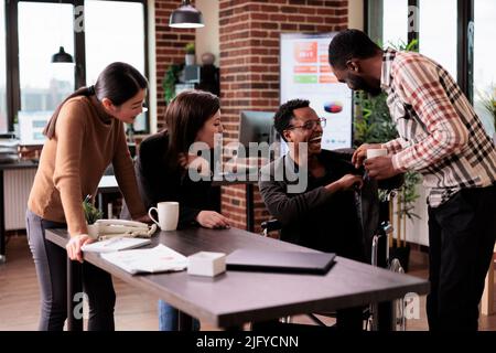 Uomo afroamericano con disabilità fisica divertirsi al lavoro, prendersi una pausa con i compagni di lavoro. Utente di sedia a rotelle maschio che soffre di invalidità cronica ridendo con i partner commerciali. Foto Stock