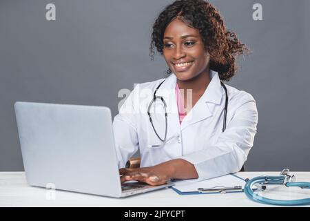 Medico donna africana in un cappotto bianco e si siede a un tavolo in un ufficio medico e lavora su un computer, telemedicina, consultazione paziente online Foto Stock
