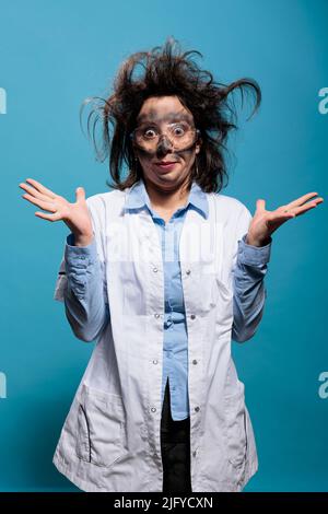 Scienziato pazzo insano e sciocco con acconciatura sudicia e faccia sporca non capisce dove l'esperimento è andato storto. Chimico MAD con espressione goofy in piedi su sfondo blu. Foto studio Foto Stock