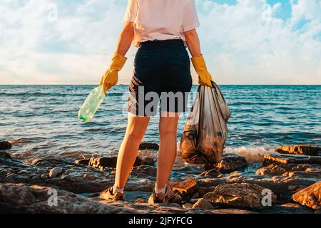 Una donna si alza con una borsa di plastica e una bottiglia nelle sue mani. Vista posteriore. Primo piano delle gambe. Spazio di copia. Pulizia della zona costiera. Il concetto di Terra Foto Stock