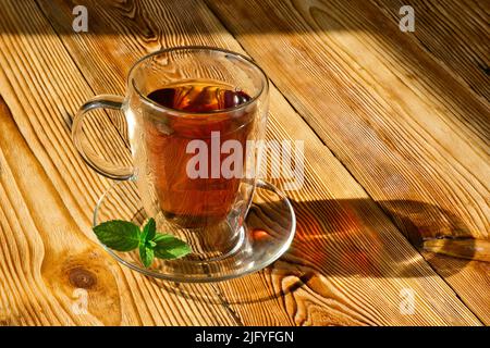 Tazza di vetro trasparente di tè nero caldo con foglie di menta fresca su tavola di legno. Foto Stock