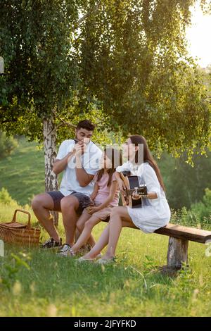 ragazza felice seduta su panchina sotto la betulla vicino ai genitori che suonano armonica e chitarra Foto Stock