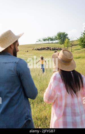 vista posteriore di coltivatori sfocati in cappelli di paglia guardando la figlia che corre verso pascolo mandria Foto Stock