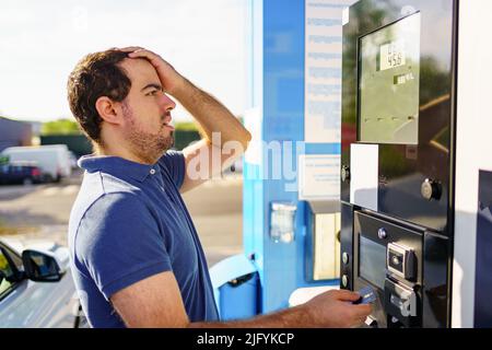 Giovane caucasico sorpreso dai prezzi elevati del carburante sul quadro di valutazione della stazione di servizio. Foto Stock
