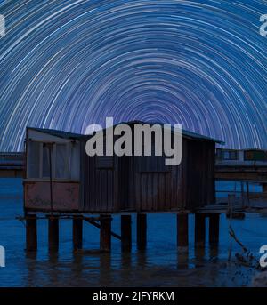 Percorsi stellari nel cielo notturno in Italia. Movimento delle stelle dietro un vecchio capannone di legno e metallo in piedi in acqua Foto Stock