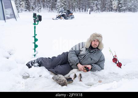 Uomo giacente su ghiaccio mentre pescano da un buco, Lapponia, Svezia, Scandinavia, Europa Foto Stock