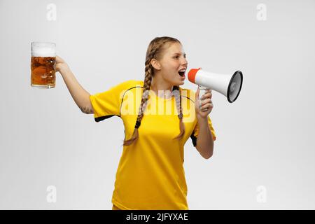 Giovane donna eccitata in divisa da calcio gialla con tazza di birra e supporta la squadra preferita. Appassionati di calcio, competizione, sport, concetto oktoberfest Foto Stock