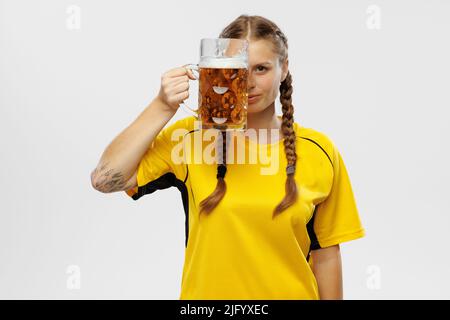 Una giovane donna eccitata in una divisa da calcio gialla che guarda attraverso la tazza di birra e supporta la squadra preferita. Appassionati di calcio, competizione, sport, oktoberfest Foto Stock