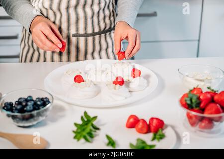 Cucina, facendo meringa fatta in casa. Donne mani che decorano torte Pavlova con frutti di bosco freschi sul tavolo da cucina con ingredienti. Ricette per delicio Foto Stock