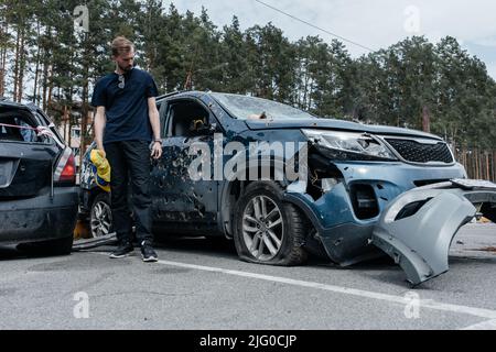 L'uomo si alza vicino a un'auto danneggiata sparata con proiettili. Auto fucilata dall'esercito russo durante l'evacuazione dei civili. Cesoie e fori a pallottola Foto Stock