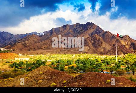 Hatta City segno di benvenuto scritto con grandi lettere poste in montagne Hajar e bandiera degli Emirati Arabi Uniti volare alto in Hatta enclave di Dubai negli Emirati Arabi Uniti Foto Stock