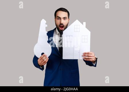 Shocked uomo porticato che tiene la casa di carta e la chiave grande, acquisto del bene immobile, servizi di affitto, ipoteca, vestendo il vestito ufficiale di stile. Studio interno girato isolato su sfondo grigio. Foto Stock