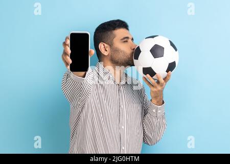 Ritratto di bell'uomo bearded in piedi baciare la palla di calcio e mostrando il telefono cellulare con display nero vuoto, indossando camicia a righe. Studio interno girato isolato su sfondo blu. Foto Stock