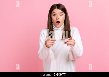 Donna estremamente stupita che punta alla macchina fotografica, essendo molto scioccata per scegliervi, mantiene la bocca aperta, indossando il maglione casual bianco di stile. Studio interno girato isolato su sfondo rosa. Foto Stock