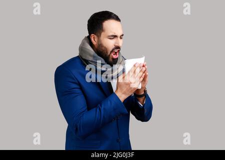 Vista laterale dell'uomo avvolto in caldo sciarpa starnuti in tovagliolo, pulizia naso che cola, dolore allergico o sintomi influenzali, epidemia stagionale di influenza. Studio interno girato isolato su sfondo grigio. Foto Stock