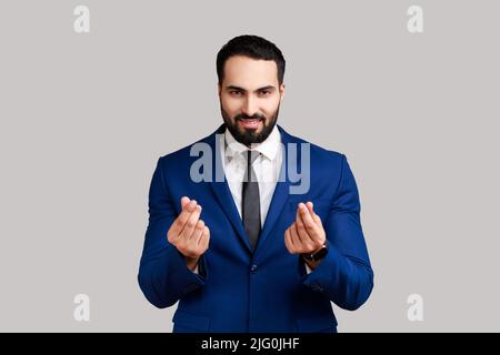 Ritratto di bel businessman barbuto sorridendo slyly e facendo il gesto di soldi, chiedendo il pagamento, l'indennità, indossando il vestito ufficiale di stile. Studio interno girato isolato su sfondo grigio. Foto Stock