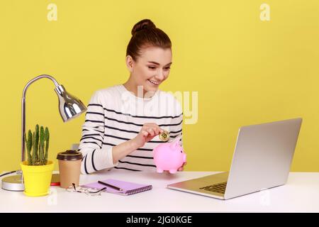 Ritratto di felice sorridente donna dai capelli scuri investendo in bitcoins, mettendo la moneta di cripto dorato in banca di piggy e guardando il display del portatile. Studio interno girato isolato su sfondo giallo. Foto Stock