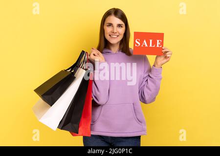 Felice donna felice che tiene in mani borse di shopping di carta e carta di vendita, guardando la macchina fotografica con piacevole sorriso, indossando felpa con cappuccio viola. Studio interno girato isolato su sfondo giallo. Foto Stock