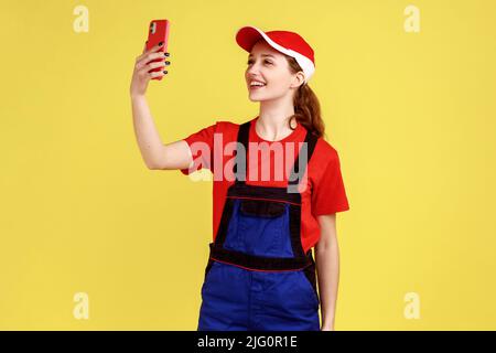 Ritratto di donna lavoratore soddisfatto in piedi con il telefono cellulare in mano e prendere selfie o trasmissione in live streaming, supporto online. Studio interno girato isolato su sfondo giallo. Foto Stock