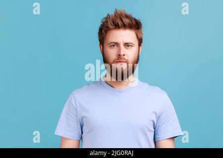 Ritratto di uomo fiducioso stretto barbuto guardando direttamente la macchina fotografica con serio volto attento, ascoltando attentamente, unsmiling determinato maschio. Studio interno girato isolato su sfondo blu. Foto Stock