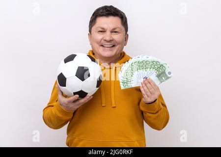 Uomo sorridente di mezza età che mostra calcio palla e divertimento di centinaia di euro fatture, vincendo molto denaro scommesse per lo sport, indossando felpa con cappuccio urban style. Studio interno girato isolato su sfondo bianco. Foto Stock