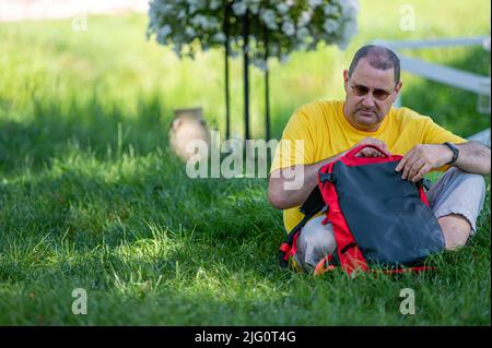 un uomo di mezza età con uno zaino siede sull'erba in un parco in una giornata di sole Foto Stock