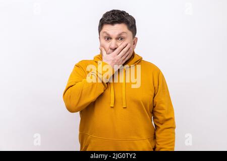 Non dirò nessuno. Ritratto di uomo confuso che copre la bocca con la mano, mantenendo terribile verità segreta, non vuole parlare, indossando una felpa con cappuccio in stile urbano. Studio interno girato isolato su sfondo bianco. Foto Stock