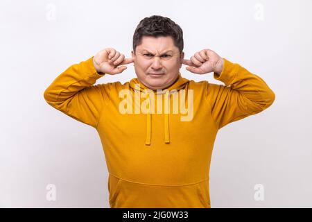 Non mi sento. Ritratto di uomo irritato dal rumore che copre le orecchie per evitare fastidiosi discorsi, non vuole ascoltare, indossando una felpa con cappuccio urban style. Studio interno girato isolato su sfondo bianco. Foto Stock