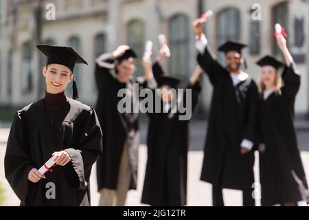Sorridente bachelor in cap tenendo diploma vicino blurred gli amici interrazziali all'aperto Foto Stock